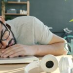 ‘스트레스 리커버리’ 시대 — 번아웃을 예방하는 5분 루틴 Man sleeping at desk with headphones and coffee.